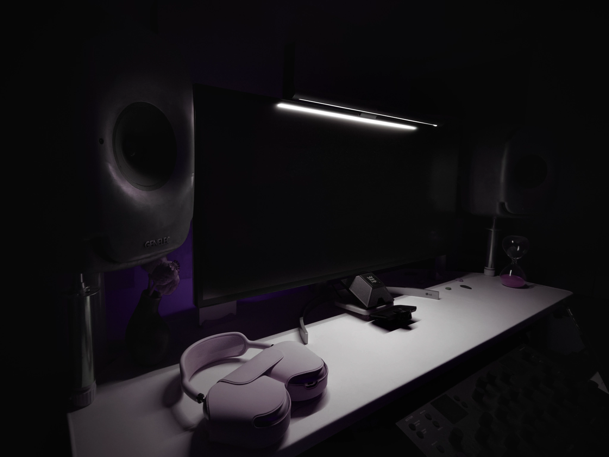 Angled view of the studio desk at night with a Genelec monitor, wide screen and headphones under a light bar